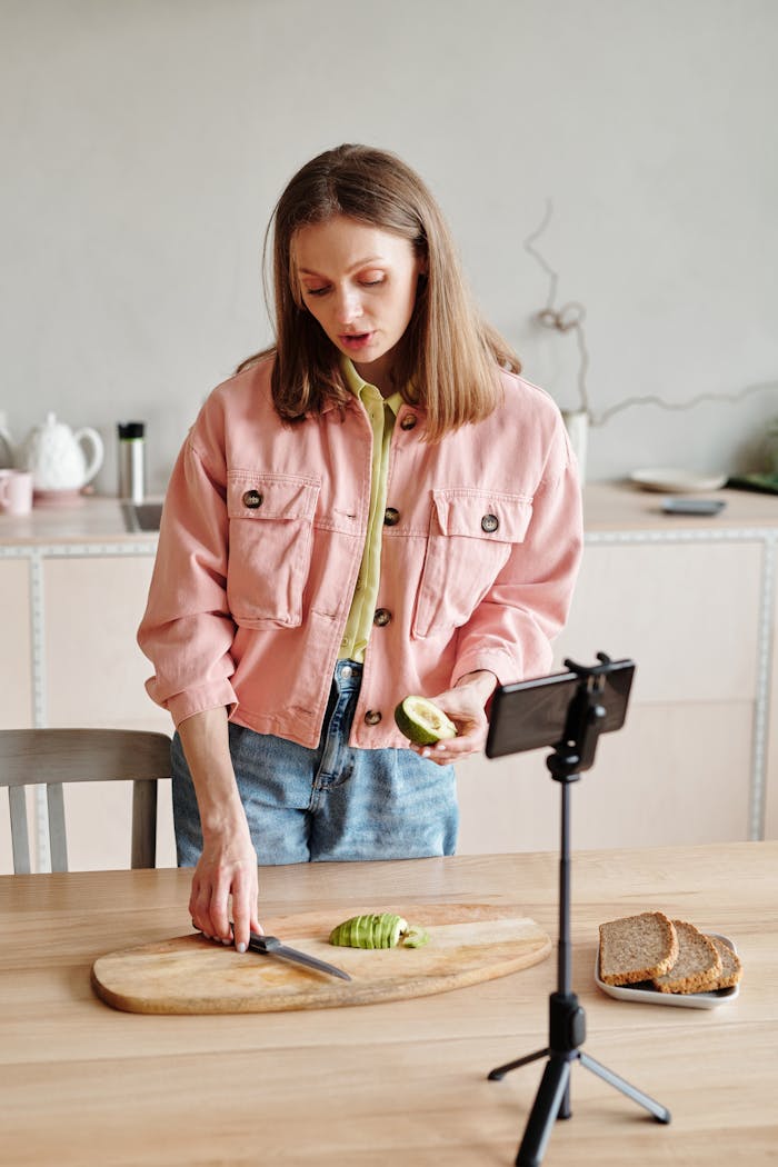 Contact Woman slicing avocado while recording video in kitchen with smartphone on tripod.