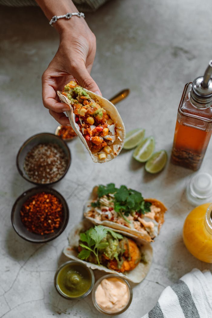 Services Mouthwatering tacos with toppings held by hand, surrounded by Mexican ingredients on a table.